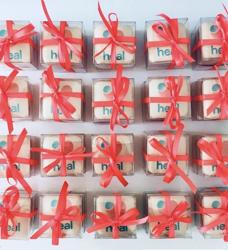 A grid of white favour boxes tied with red ribbon, each holding a square macaron printed with the Heal logo